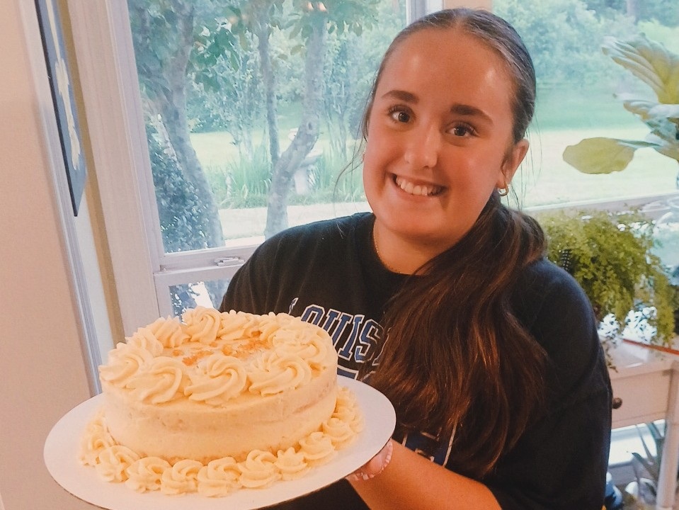 Lelia smiling and holding a cream-frosted cake by a sunny window