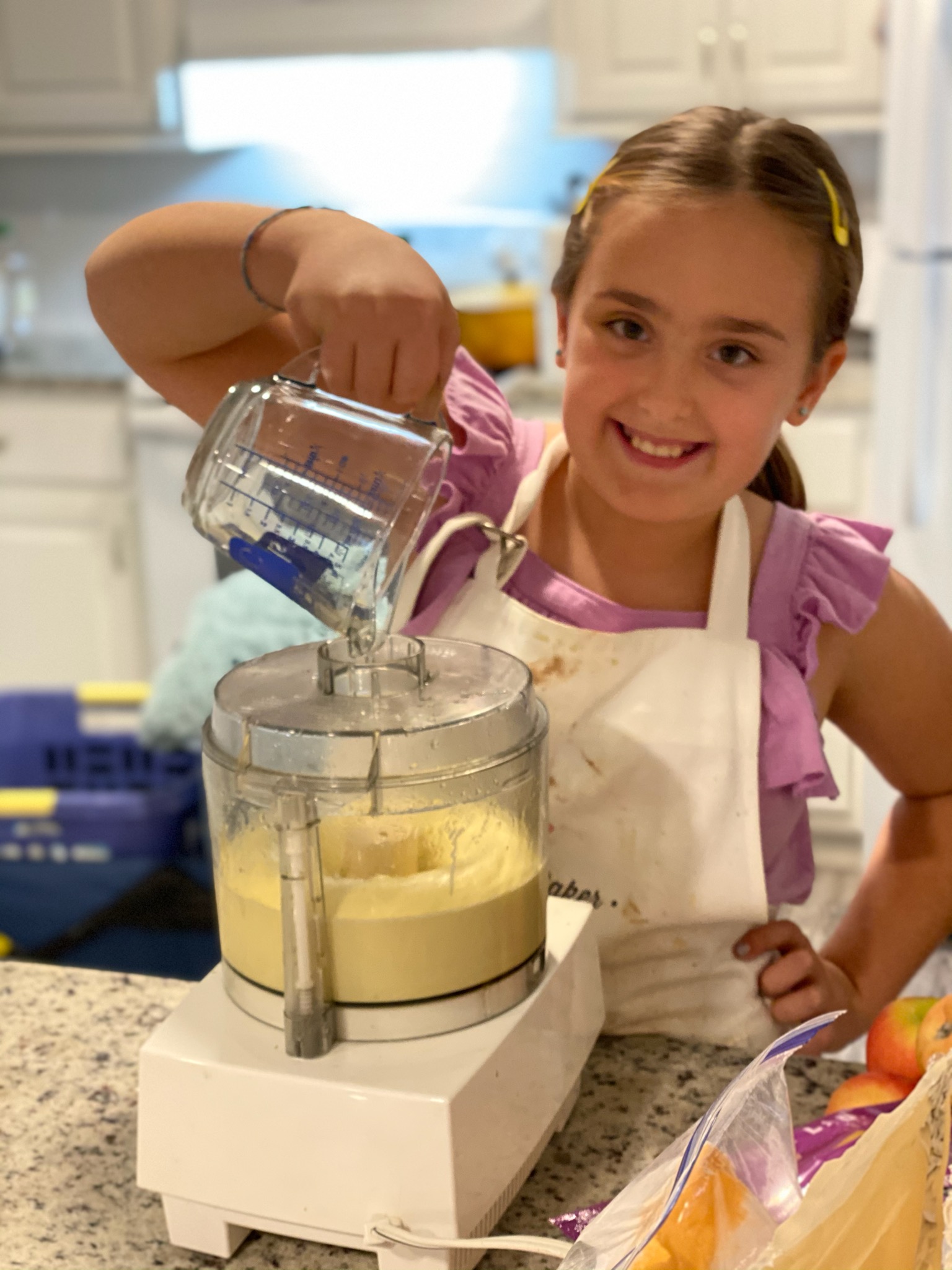 Little Lelia in her baking apron, smiling as she pours into the mixer